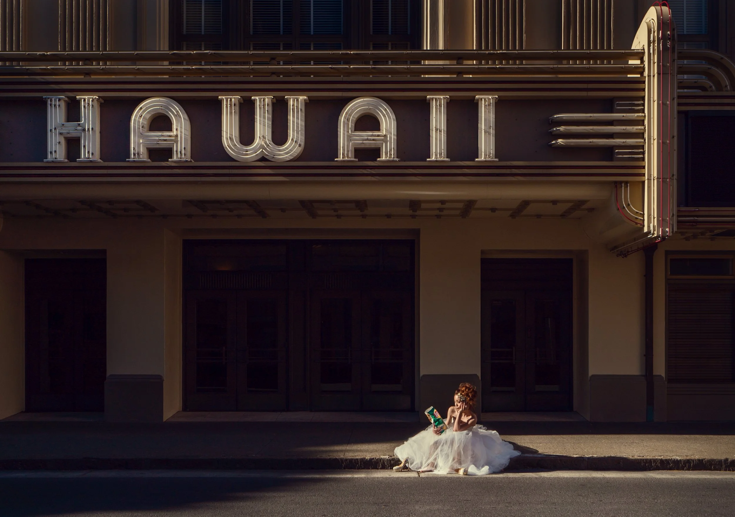 A woman in a white dress sitting outside a building with a large neon 'HAWAII' sign, reading a book under warm sunlight.