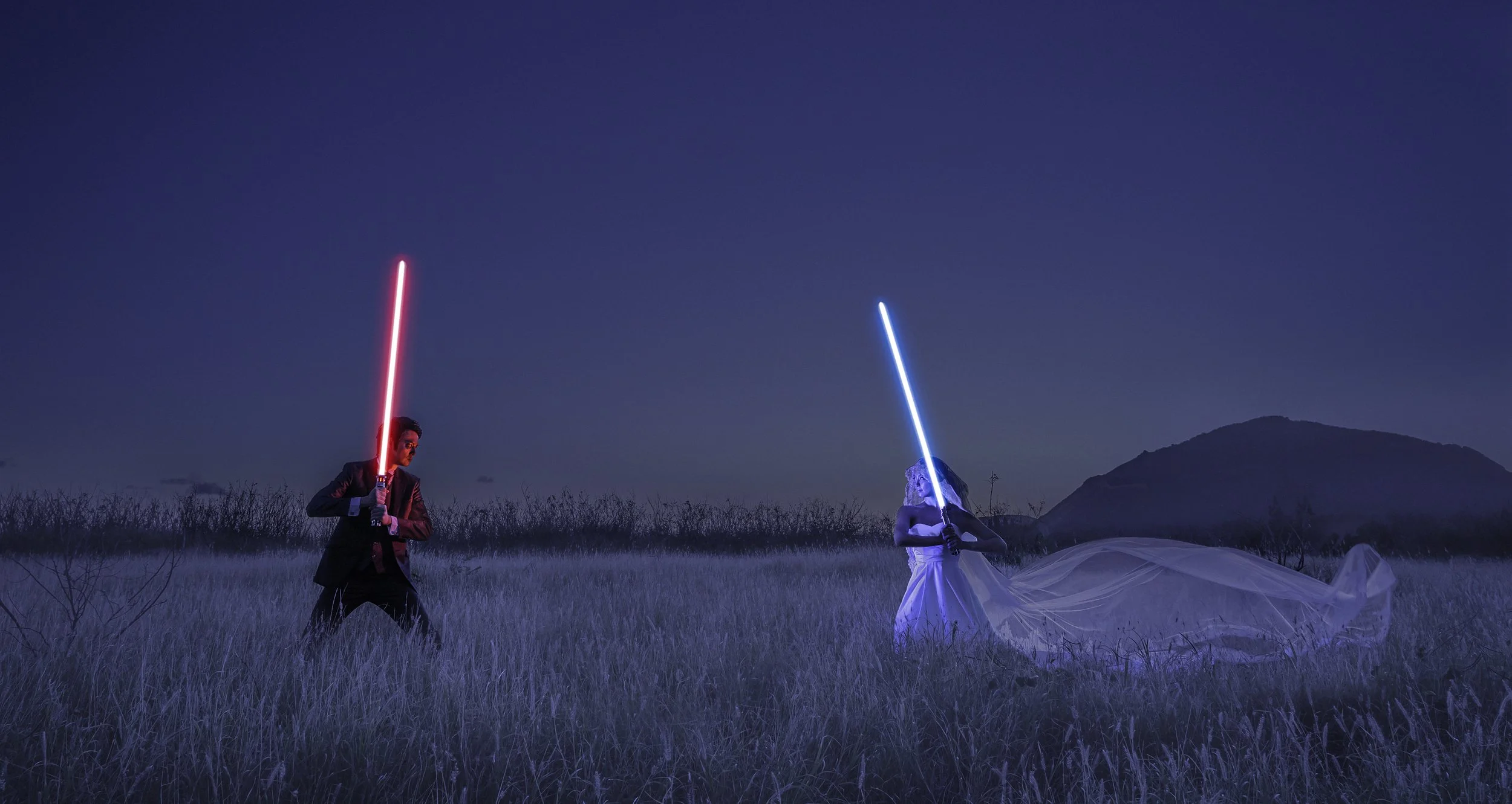 A man in a dark suit holding a red lightsaber facing a woman in a white dress with a blue lightsaber, standing in a grassy field at dusk, with a mountain in the background.