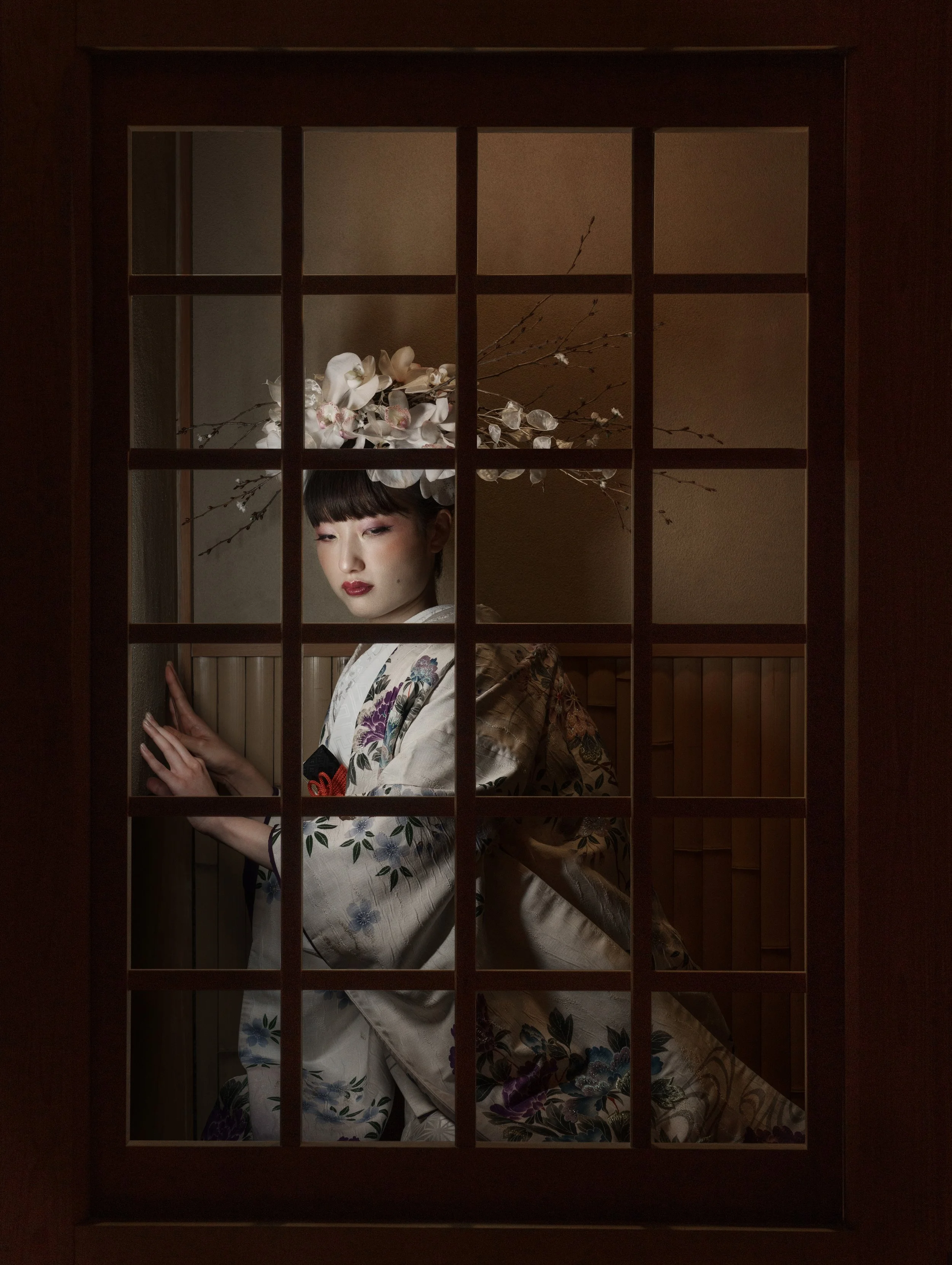 A woman dressed in traditional Japanese clothing, with a floral kimono and a headpiece of white orchids and branches, standing behind a wooden lattice door.