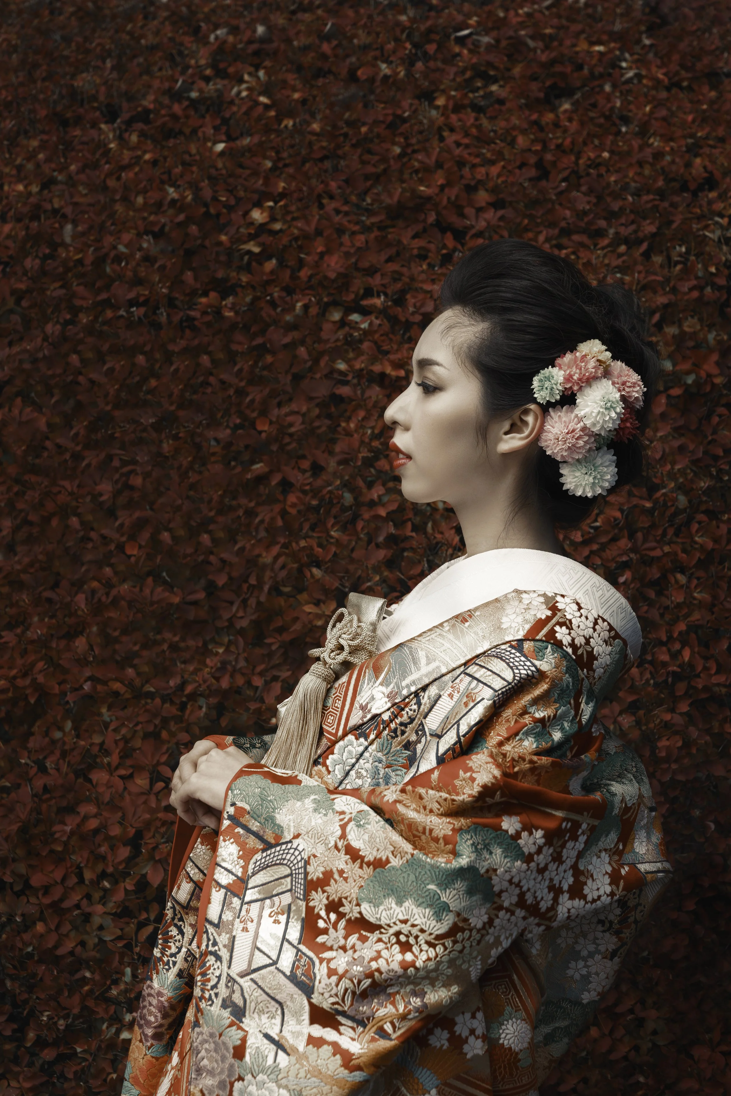 A woman wearing a traditional Japanese kimono with intricate patterns, standing in profile against a background of red foliage, with her hair styled adorned with pink, white, and green flowers.