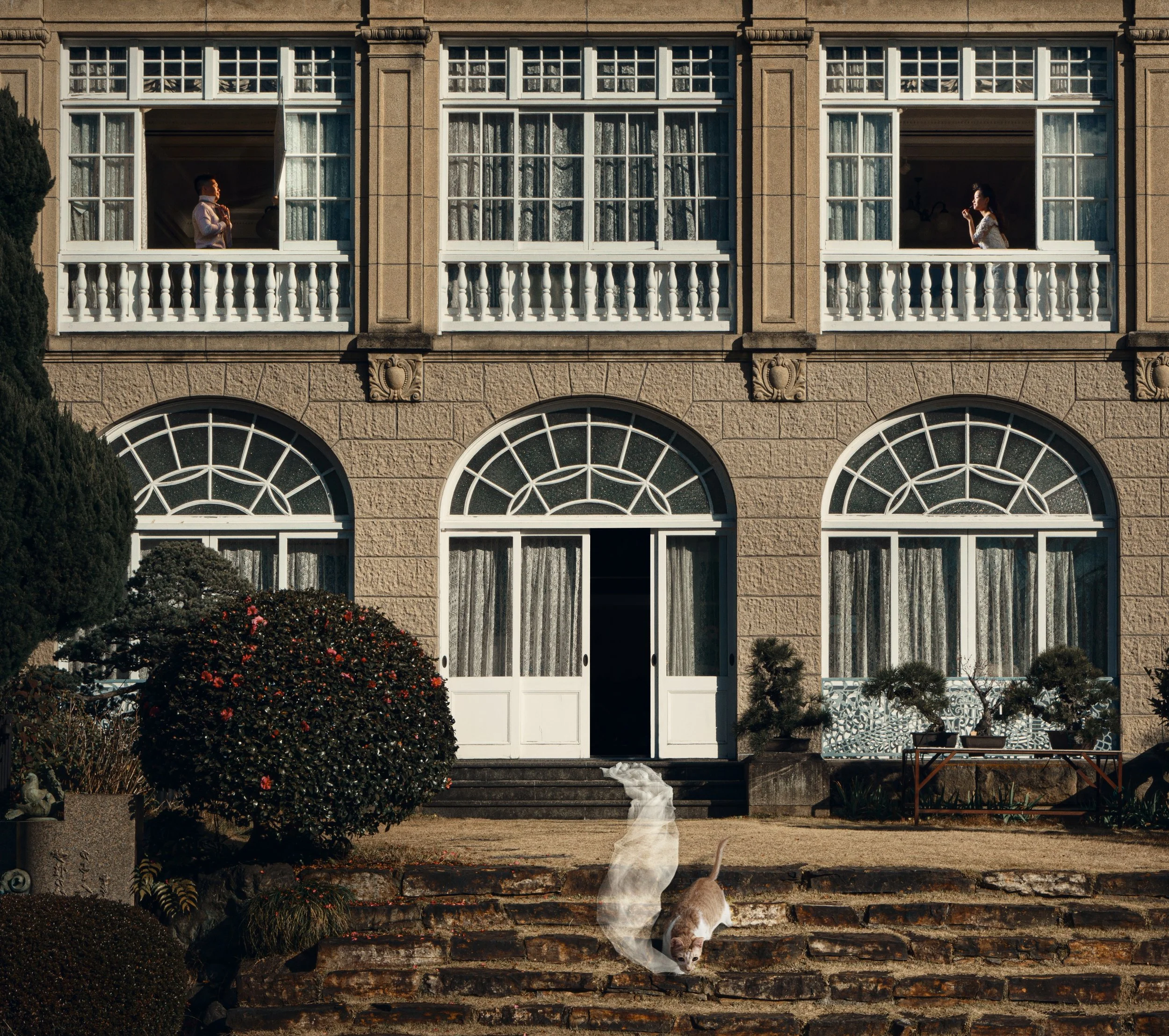 A large stone house with multiple arched windows and balconies, two women looking out from the upper windows, a cat rolling on steps in the front yard, and various bushes and plants.