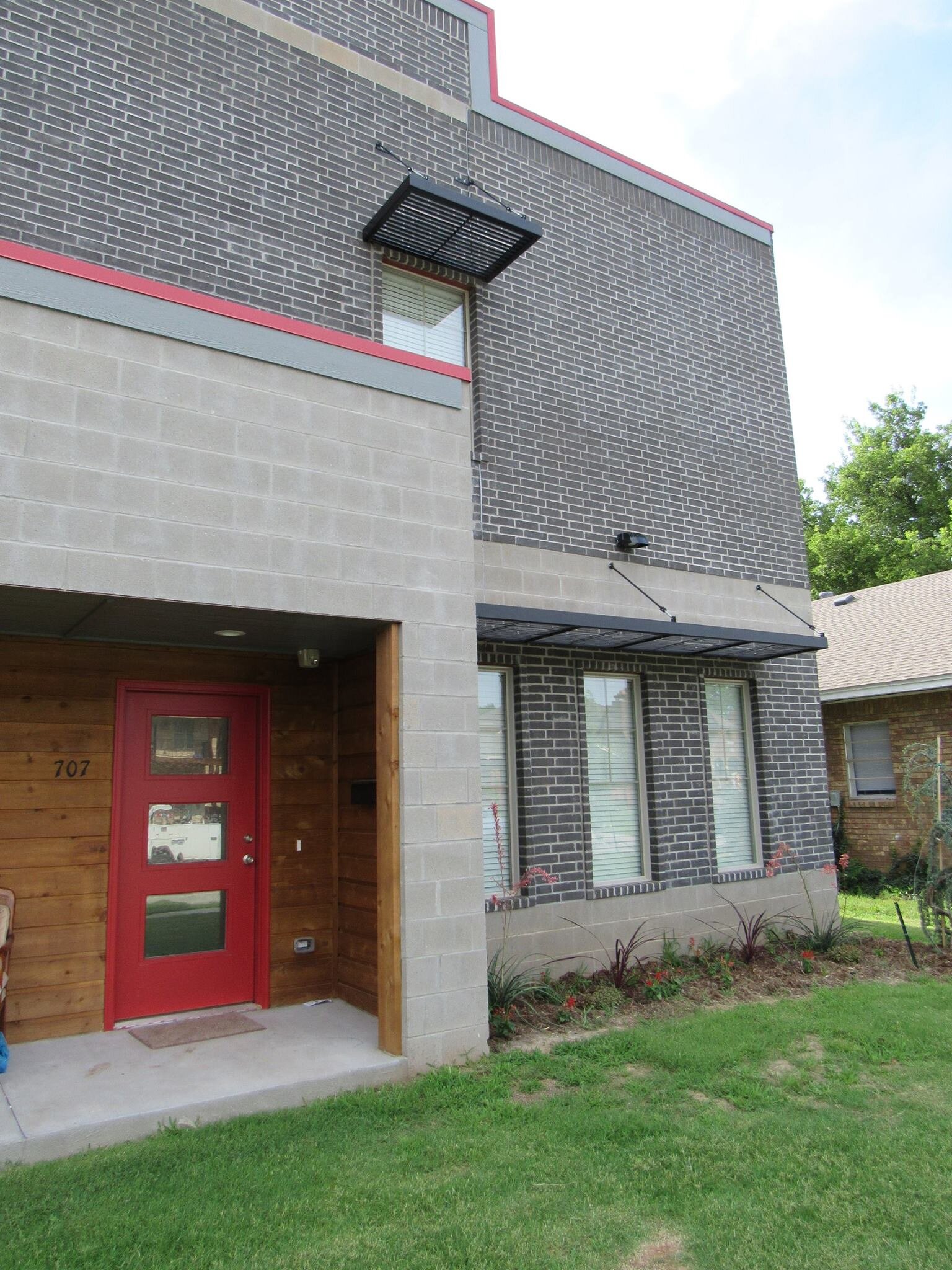 Modern two-story brick apartment building with a red door, small porch, and three windows on the lower level. The upper level features black metal awnings over the windows and a vent. The building has a mix of dark and light gray bricks and a small f