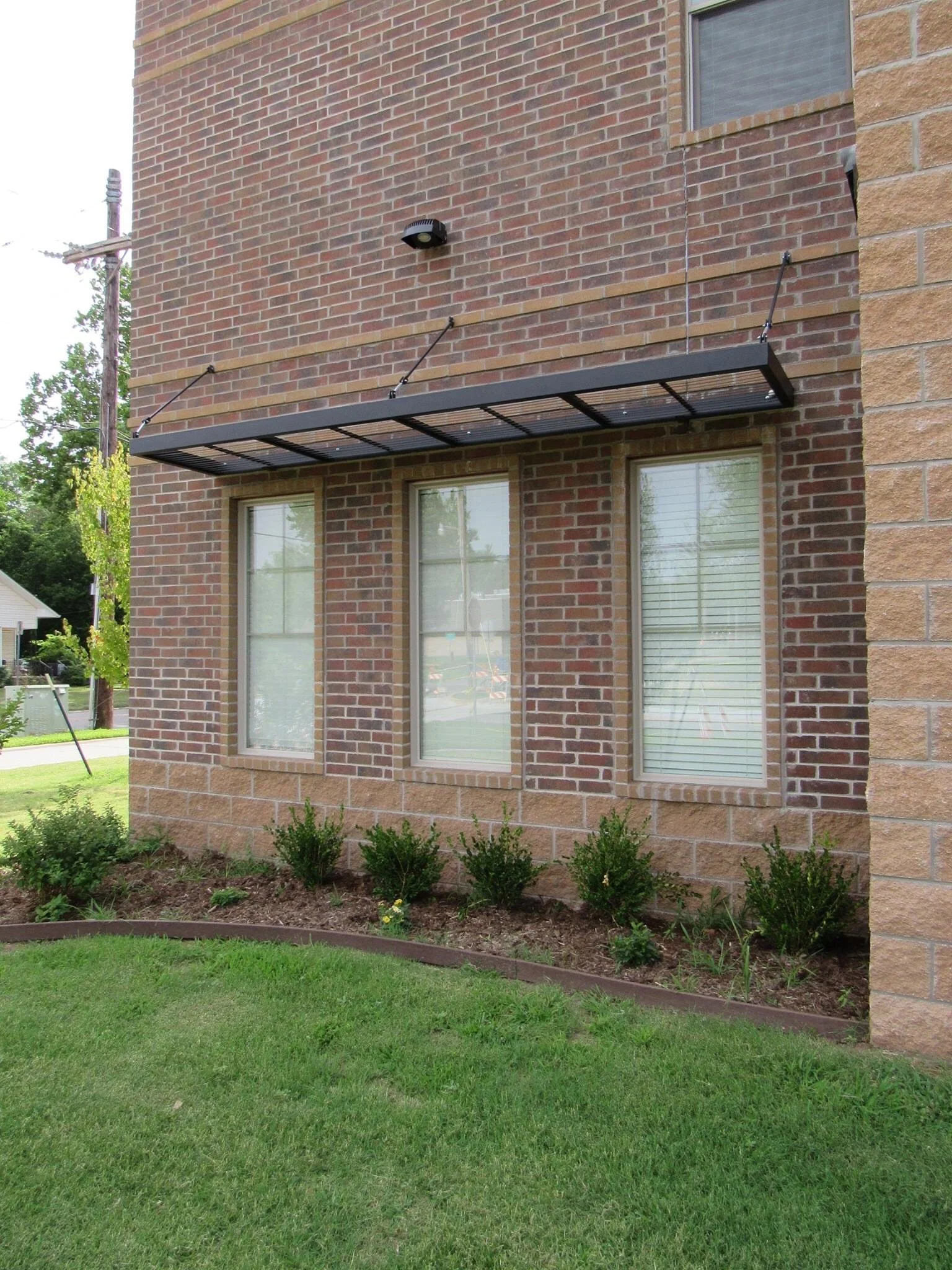 Brick building exterior with three large windows, a black metal awning above the windows, some gardening bushes below, and a grassy lawn in the foreground.