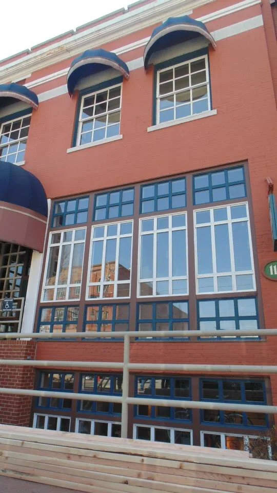 The image shows a red brick building with large multi-pane windows and decorative blue awnings above the second-story windows. There is a metal railing and a wooden barrier in the foreground.