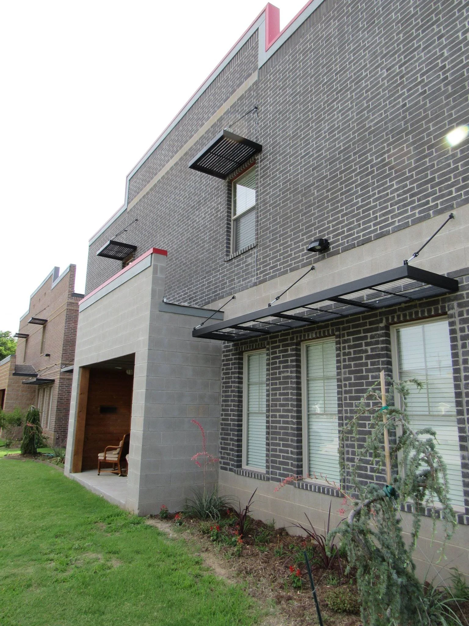 Modern townhouse with dark gray and beige brick facade, small front yard with green grass and newly planted bushes, and outdoor seating area under an overhang.