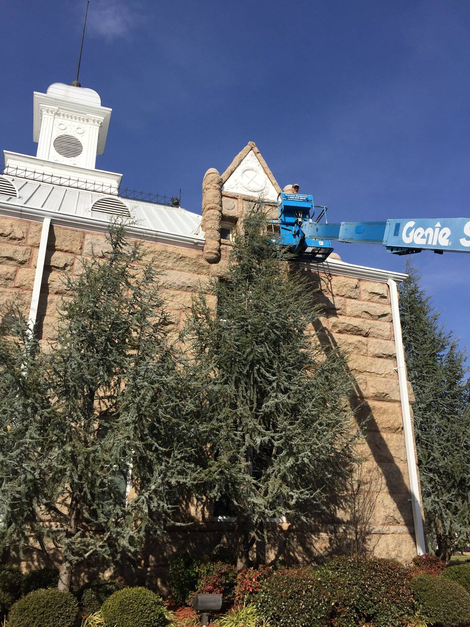 A worker on a blue lift is repairing the exterior of a stone building with a large white decorative tower against a clear blue sky. The building has trees and shrubs in the foreground.