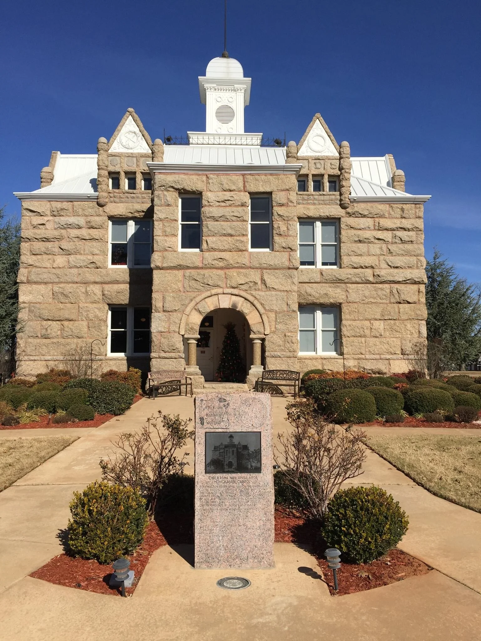 Historic stone building with a clock tower, arched entrance, and Christmas tree visible inside. Stone monument plaque with photograph and text in front of building, surrounded by small bushes and benches.