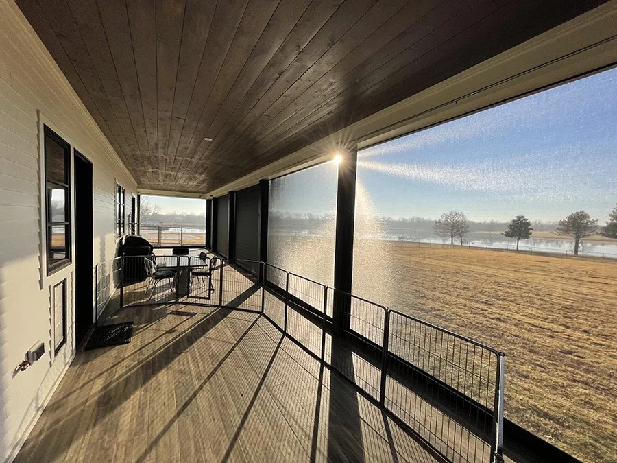 View from a porch with outdoor furniture overlooking a grassy field, trees, and a body of water in the distance during sunrise or sunset.