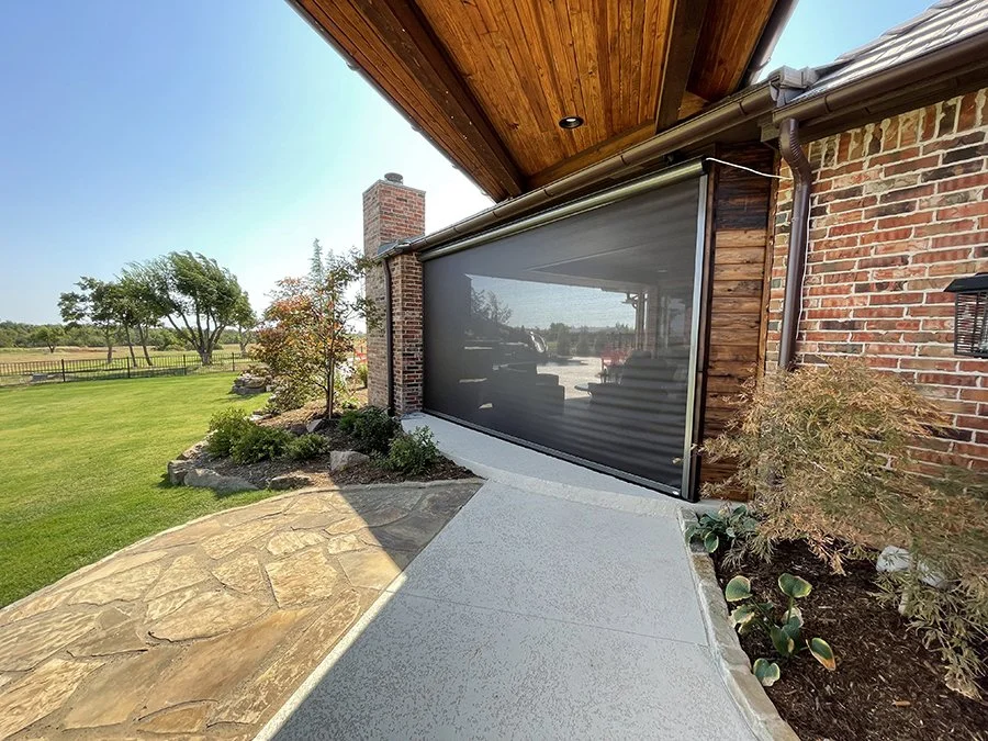 View of a house exterior with a screened porch, brick wall, stone walkway, and landscaped garden with trees and bushes.