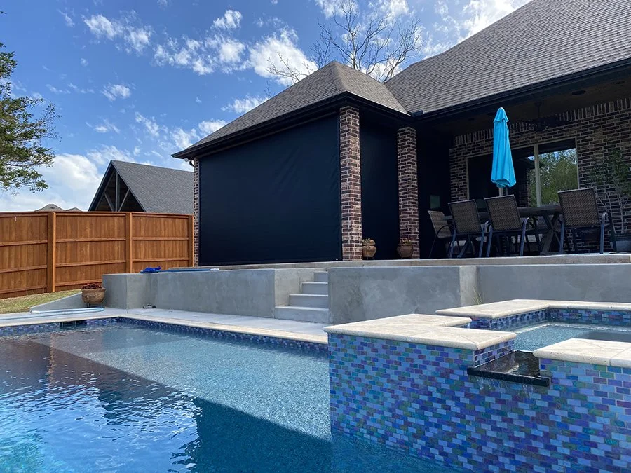 Backyard with a swimming pool, concrete deck, wooden fence, and a brick house with a patio table and umbrella.