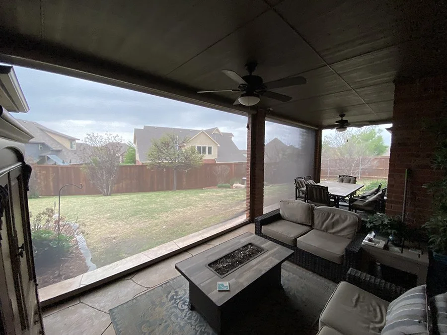 View of a backyard partially covered by an enclosed porch with furniture, ceiling fans, and a fire pit. The yard has grass, a few trees, and a wooden fence, with neighboring houses visible in the background.