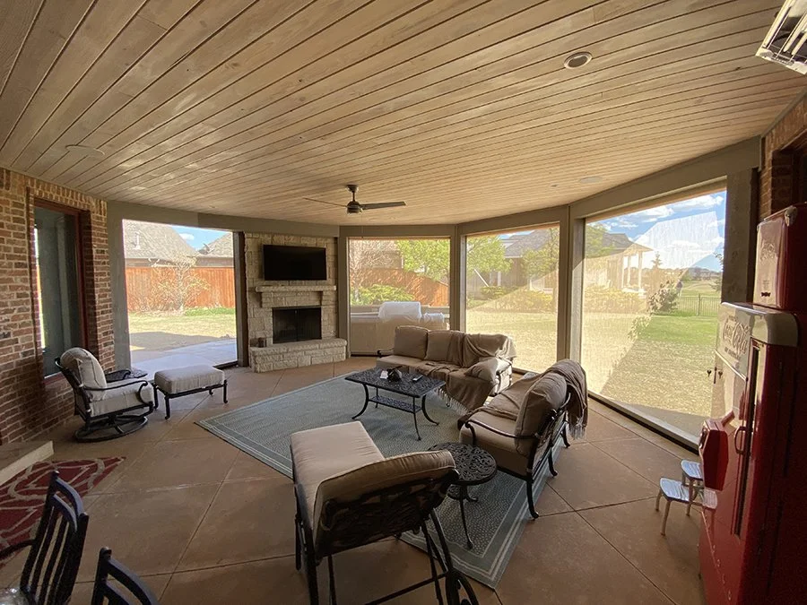 Sunlit screened porch with a seating area including a sofa, armchair, and coffee tables, brick fireplace with a mounted TV, ceiling fan, and large windows revealing a yard outside.