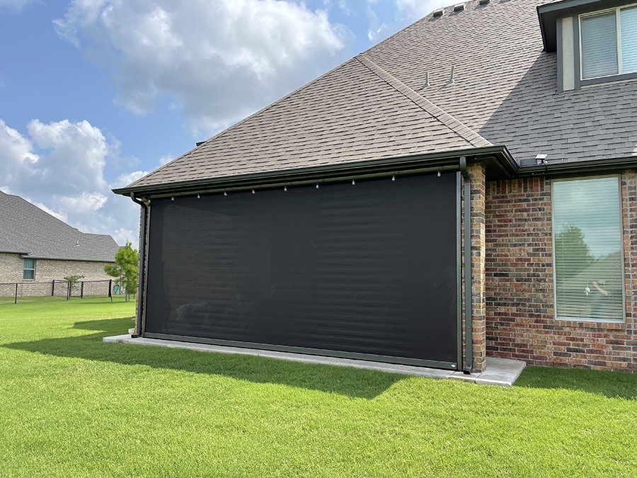 A black retractable outdoor porch or patio screen attached to a brick house, with a green lawn and neighboring houses in the background under a partly cloudy sky.
