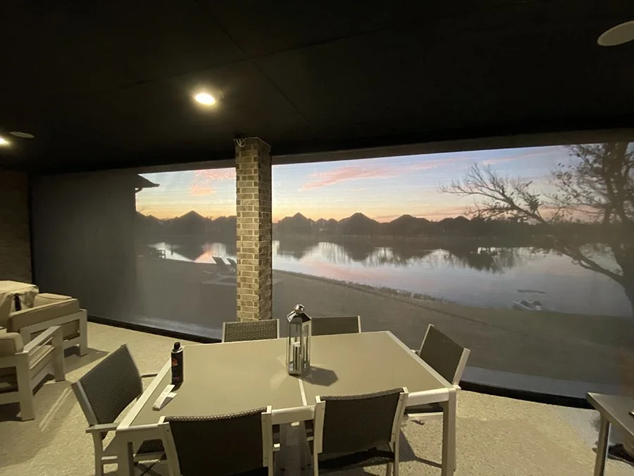 Indoor space with a large screen showing a lakeside sunset landscape, with boats on the water and trees, as furniture including a dining table, chairs, and a lantern.