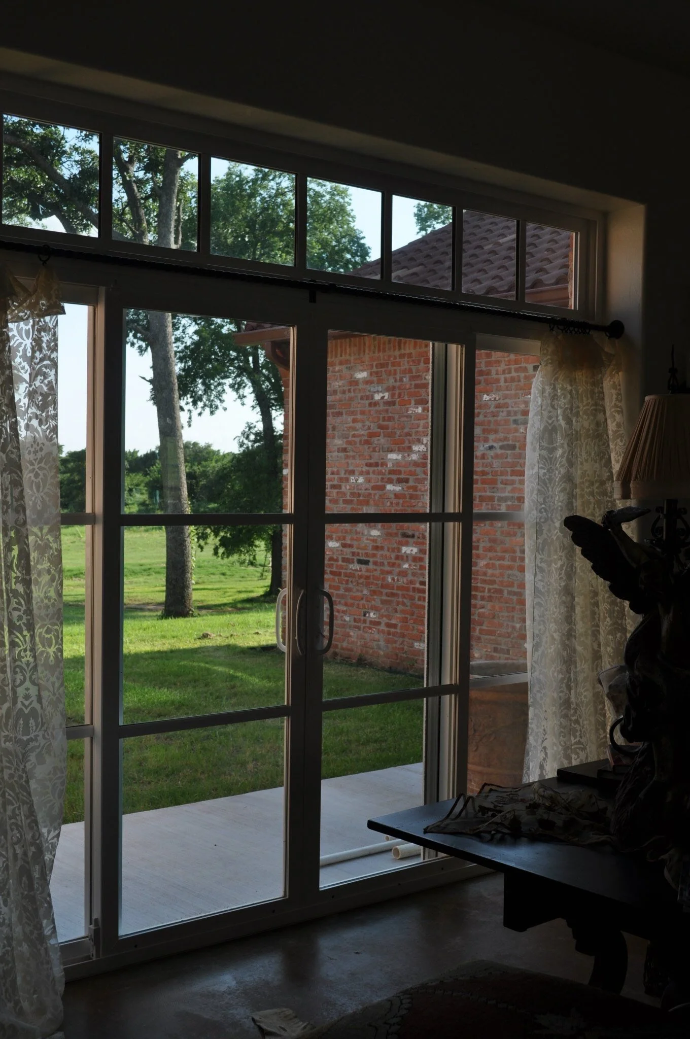 Interior view of a sliding glass door leading to a backyard with green grass, trees, and a brick wall, with lace curtains on either side.