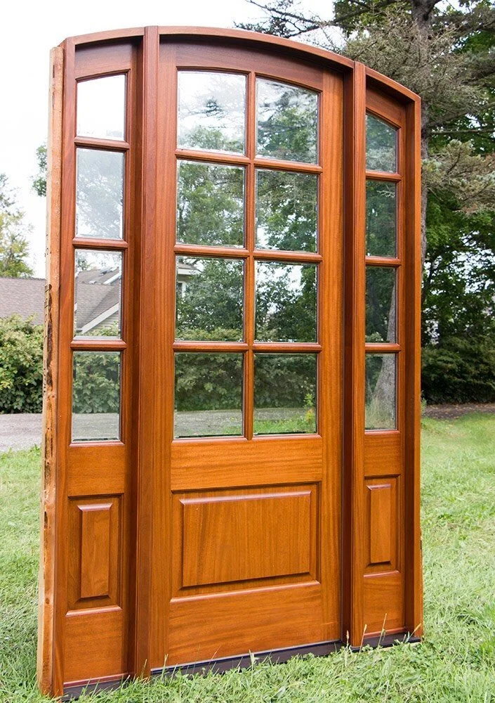 A wooden window with multiple glass panes leaning against a grassy yard with trees in the background.