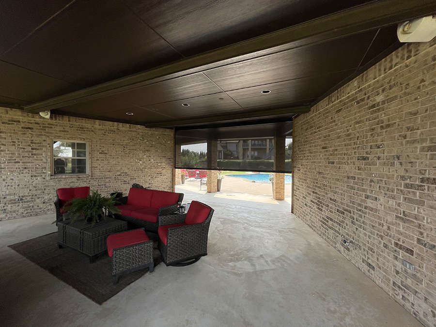 Outdoor covered patio with brick walls, ceiling, and furniture including red cushioned chairs and a sofa, with a view of a pool in the background.