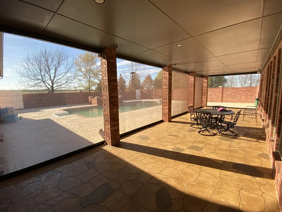 Covered patio area with stone floor, brick columns, patio furniture, and a screened enclosure overlooking a backyard with a swimming pool and trees.