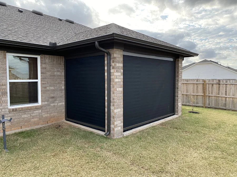 Backyard of a house with a brick exterior, showing a window and a screened porch or patio with black screens. The yard has grass, a wooden fence, and a cloudy sky.