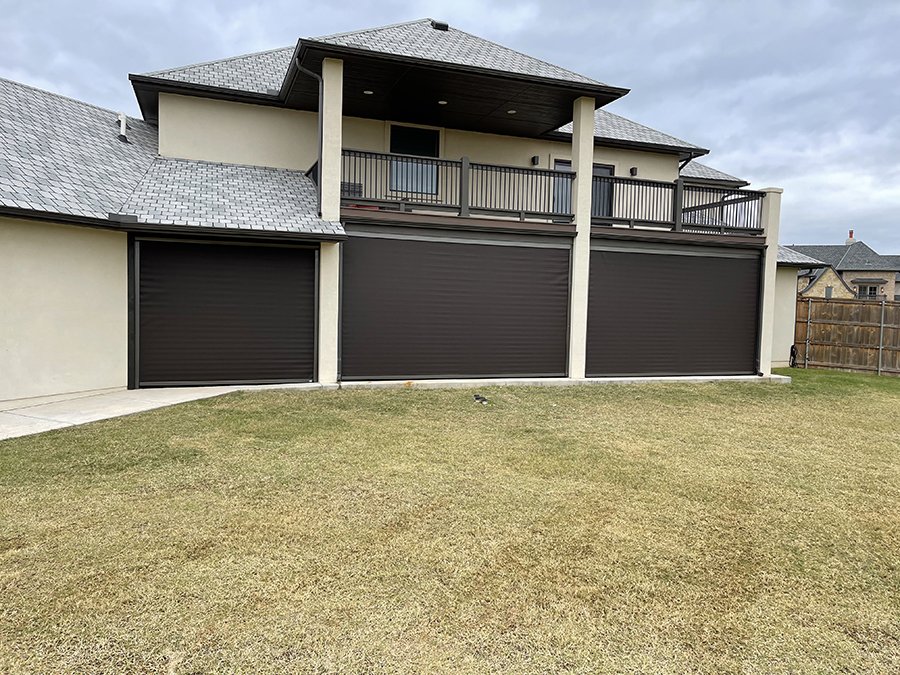 Backyard of a modern house with three black garage doors and a second-floor balcony with black railing, set against a cloudy sky.