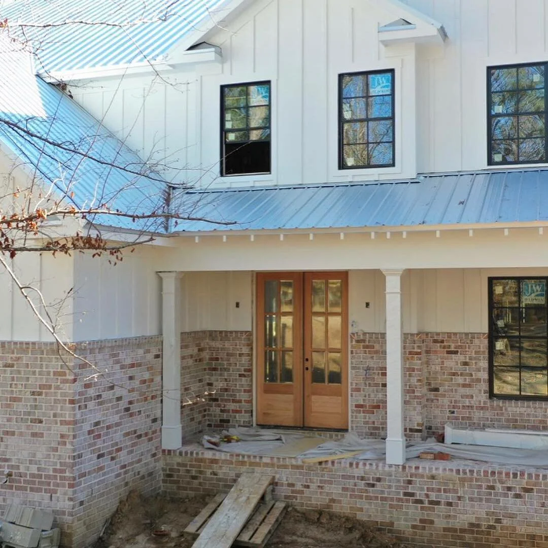 Front porch of a house under construction with a brick exterior, wooden double doors, and a metal roof, with construction materials on the ground.