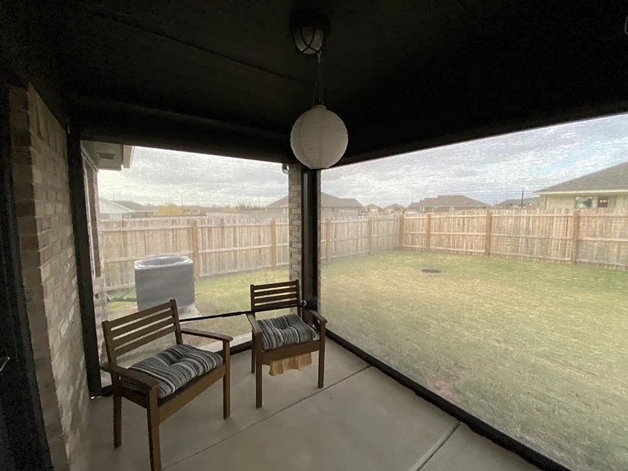 View of a backyard through a screened porch with two wooden chairs with striped cushions, a wooden side table, a black trash can, and a wooden fence around the yard.