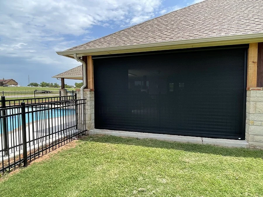 A house with a black garage door, stone pillars, green lawn, and a backyard swimming pool enclosed by a black metal fence.