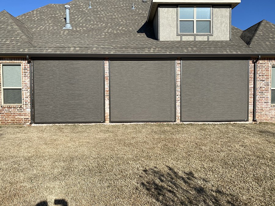 Backyard patio with three closed gray retractable screens or shades, brick exterior wall, and two windows, with a grassy yard and a clear blue sky.