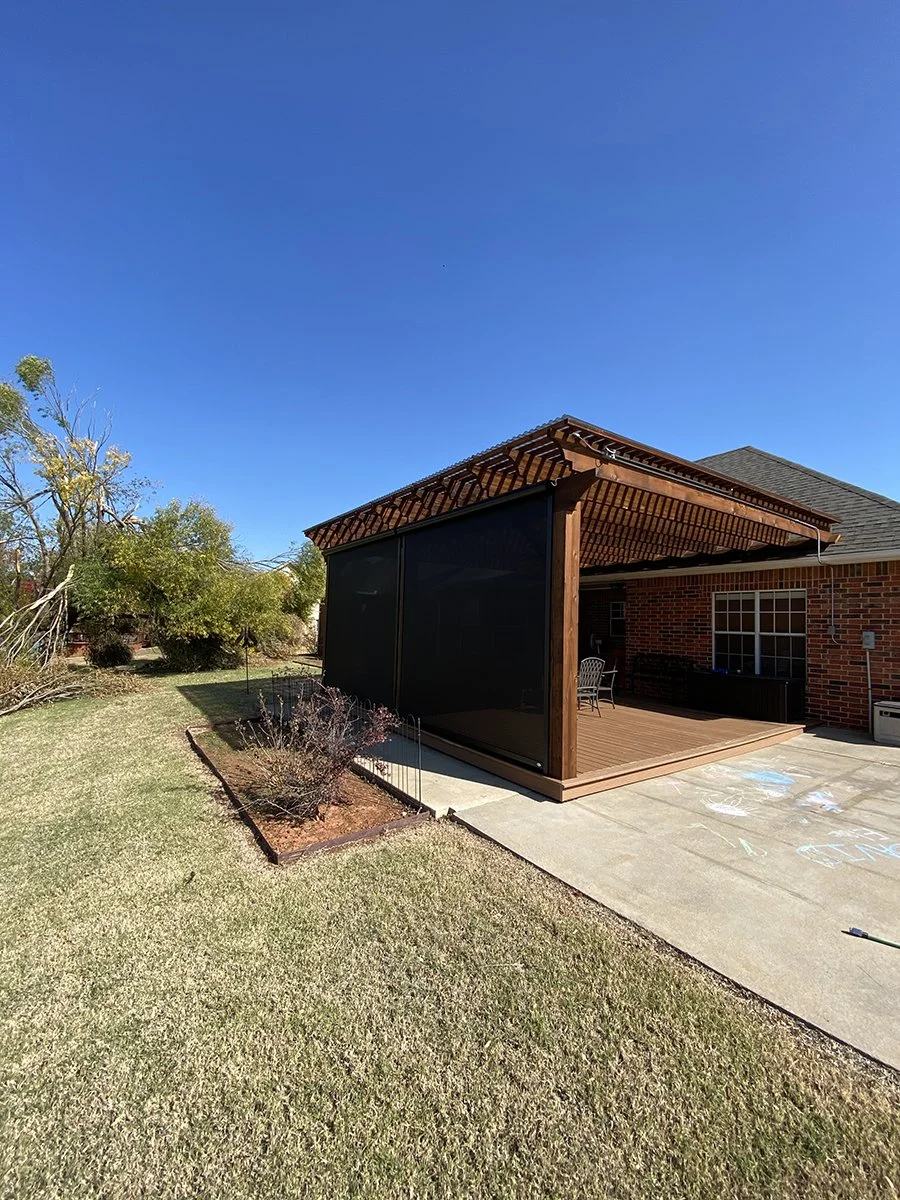 Backyard with a brick house, a wooden patio with a black screen enclosure, and a concrete patio with chalk drawings, under a clear blue sky.