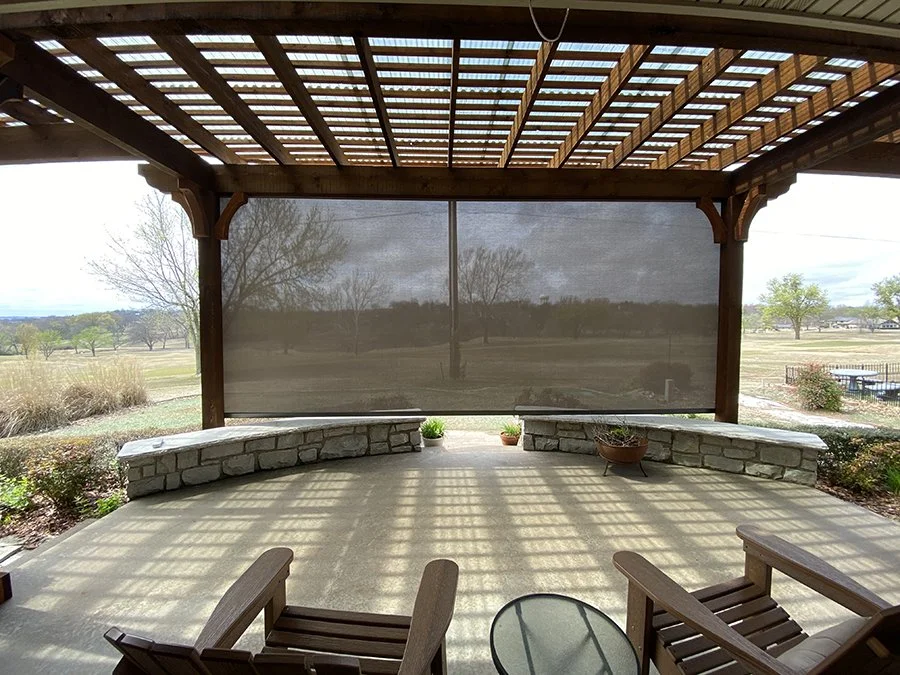 View of an outdoor covered patio with wooden pergola, stone bench, and potted plants, overlooking open grassy landscape with trees.