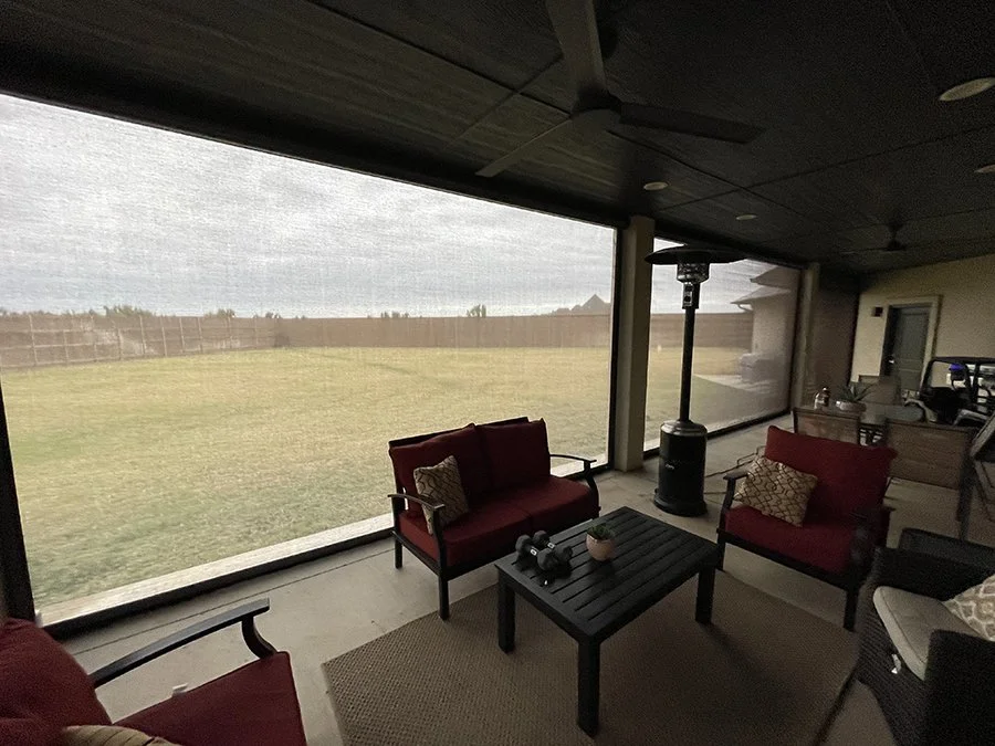 Covered patio with red outdoor sofas and beige pillows, black coffee table with binoculars and a small plant, patio heater, view through large screened windows of an open grassy yard and a wooden fence.