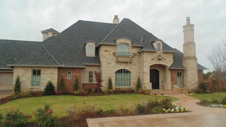 Large, elegant house with stone and brick exterior, steep dark roof, chimneys, and a small front porch with garden and bushes.