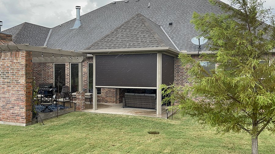 Backyard patio with a brick house, covered sitting area with chairs, a barbecue grill, and a large TV mounted under a canopy. Green grass, a young tree, and a satellite dish on the roof.