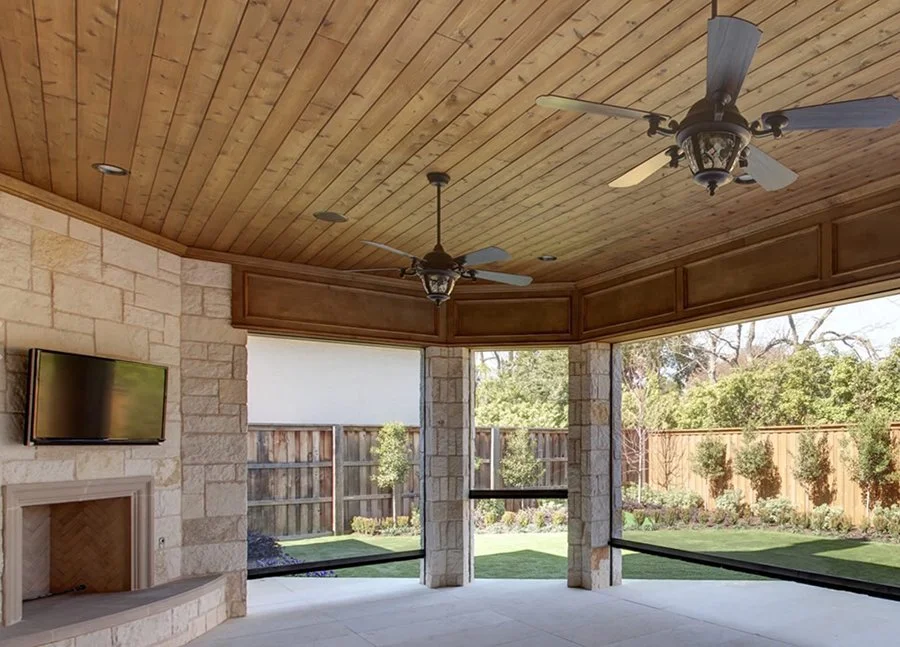 Screened backyard patio with wooden ceiling, ceiling fans, stone fireplace, and outdoor view of a fenced garden with trees and shrubs.