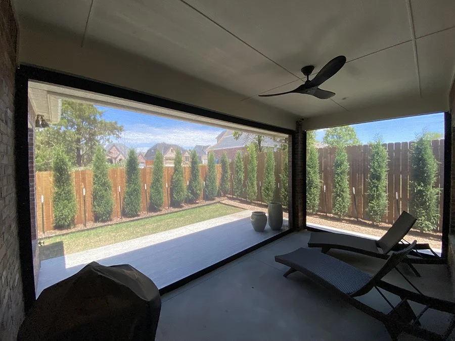 Back porch with a ceiling fan, lounge chair, potted plants, and a view of a backyard with tall green bushes and a wooden fence.