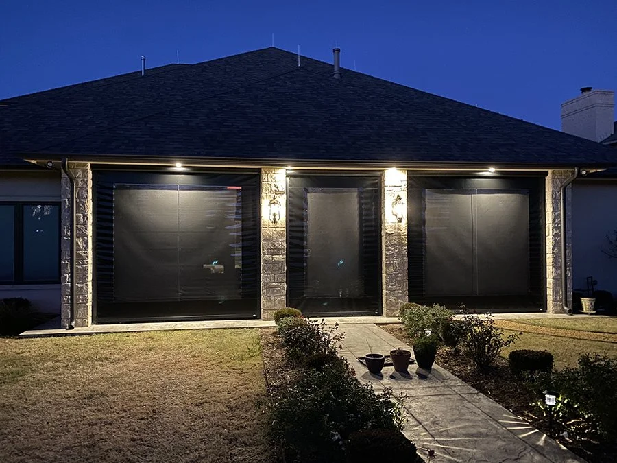 The back of a house at night with three large, closed window screens illuminated by exterior lights, a pathway leading to the house, and a small garden with potted plants and bushes.