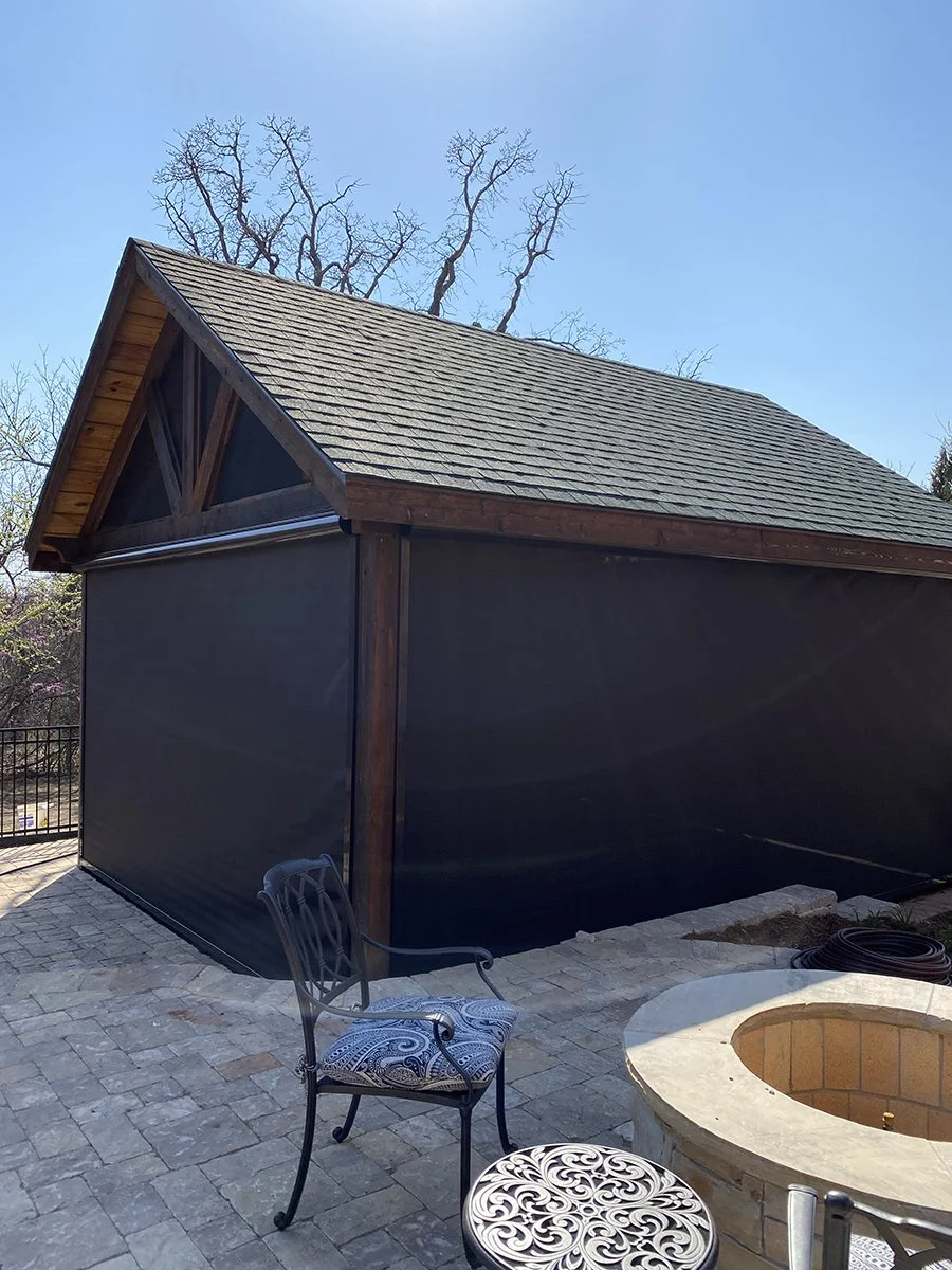 Backyard patio featuring a black privacy screen, a chair with a patterned cushion, a small ornate metal table, a fire pit, and a shed with a shingled roof and wooden trim, under a clear blue sky with some leafless trees in the background.