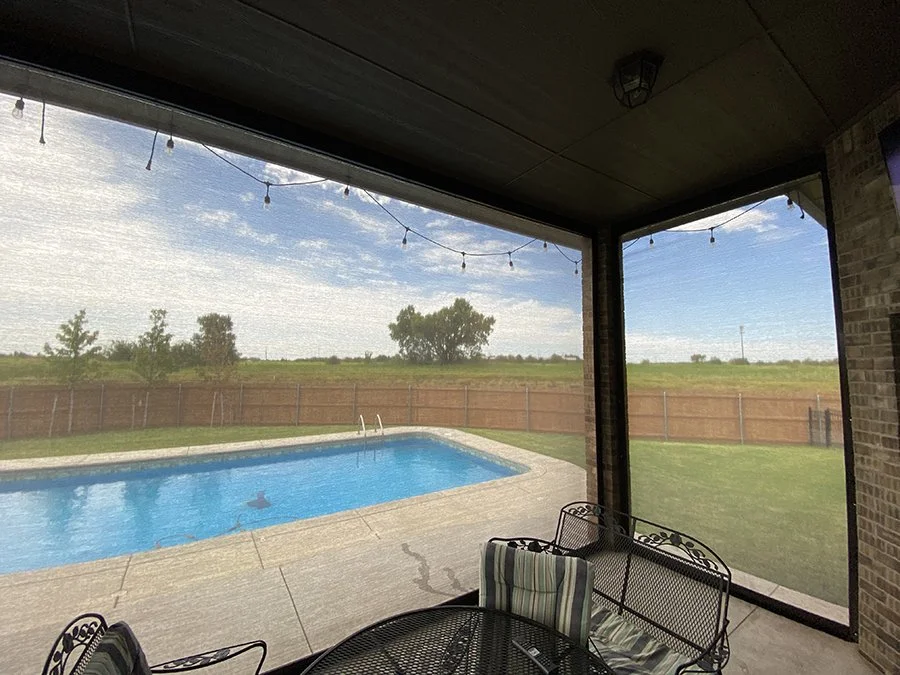 View from a screened porch showing a backyard swimming pool, a grassy area with a fence, and a sky with clouds and string lights overhead.