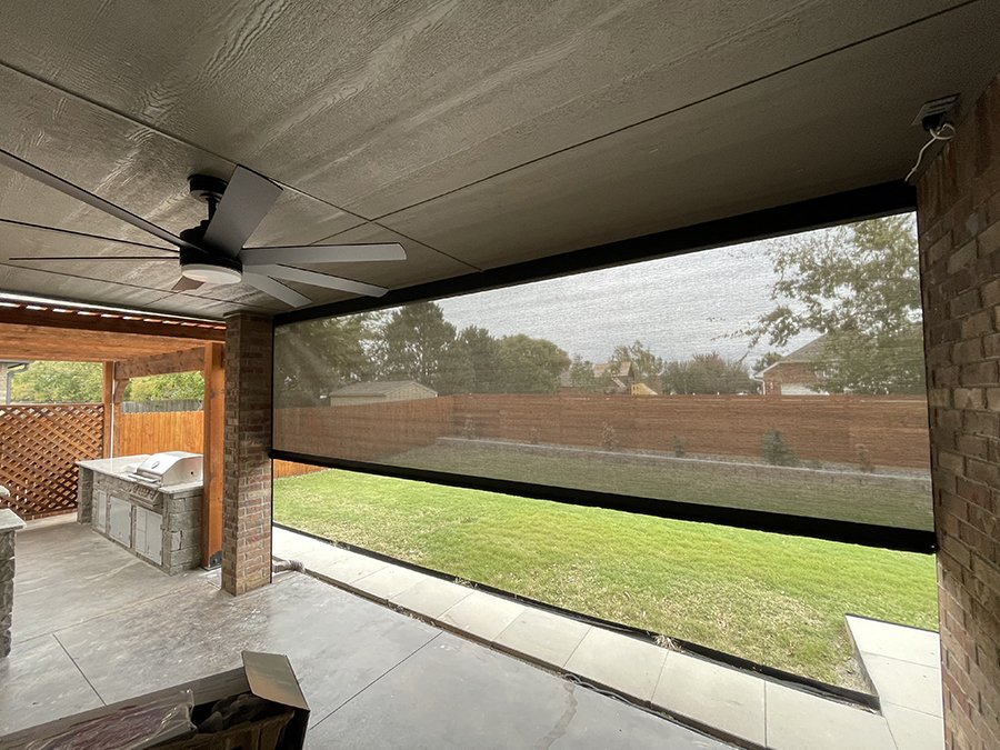 A covered outdoor patio with a ceiling fan and a mesh privacy screen, overlooking a grassy backyard enclosed by a wooden fence.