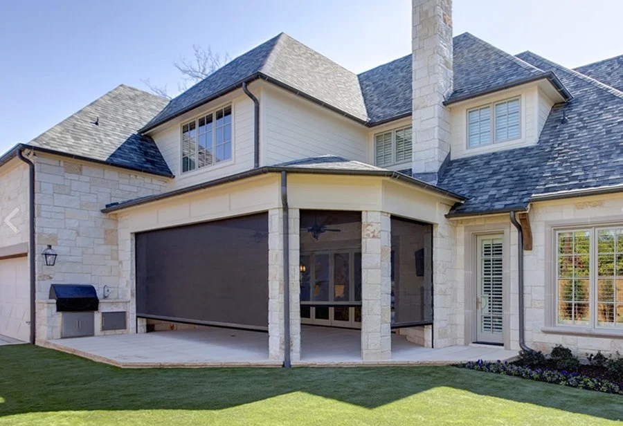 Backyard view of a large, modern house with a stone exterior, multiple gabled roofs, and a screened-in patio area with a ceiling fan.