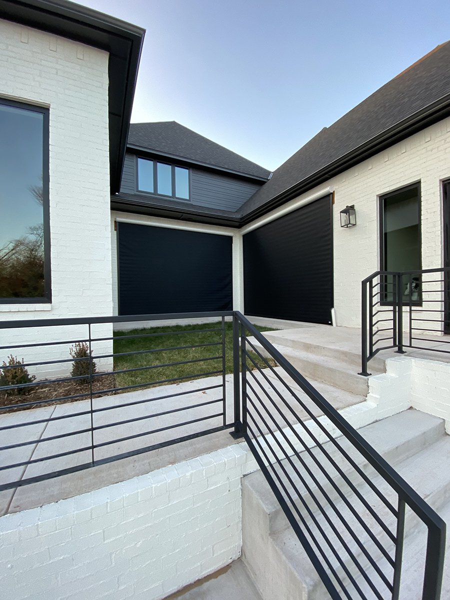 Exterior view of a modern house with black garage doors, white brick walls, and metal railings on stairs and ramp.