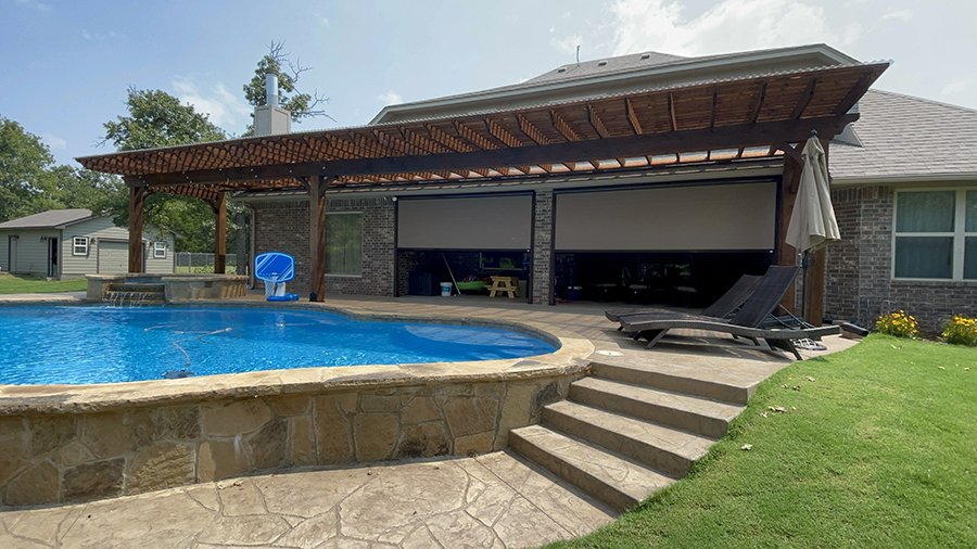 A backyard with a swimming pool, stone steps, a covered patio with a wooden roof, lounge chairs, and a house in the background.