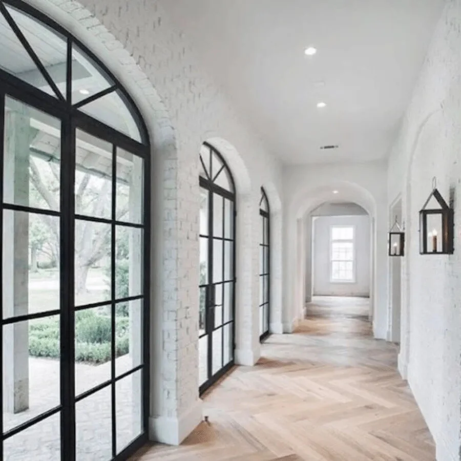 Bright hallway with white brick walls, arched windows with black frames, and light wooden herringbone flooring, featuring a minimalist decor.