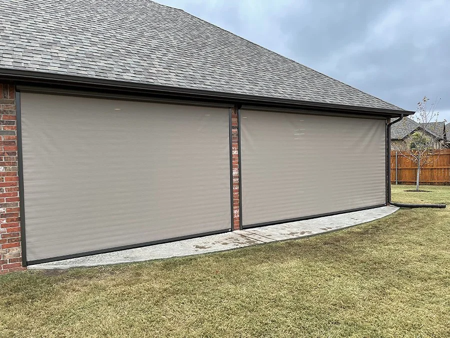 Two beige roll-up garage door shades on a brick house with a curved concrete pad and a grassy yard, with a wooden fence and trees in the background.