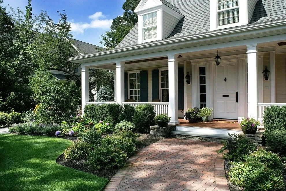 Front view of a house with a porch, white door, and black lantern lights, surrounded by green shrubs, potted plants, and a brick pathway.