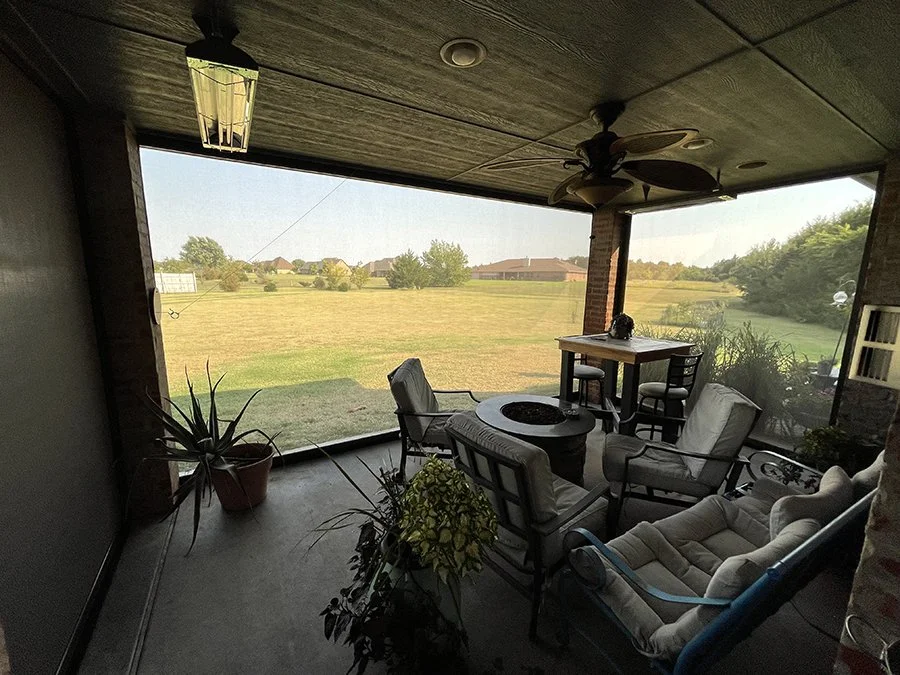 Enclosed patio overlooking a grassy field with houses in the distance, furnished with chairs, a small table, potted plants, ceiling fan, and lighting.