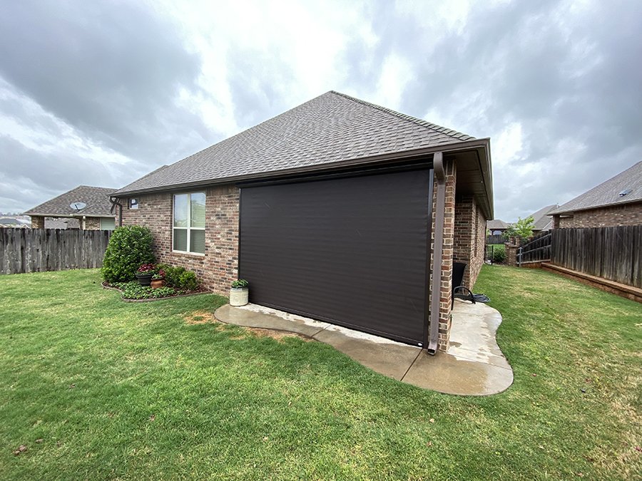 Backyard of a brick house with a black garage door, small garden with bushes, and a wooden fence, under cloudy sky.