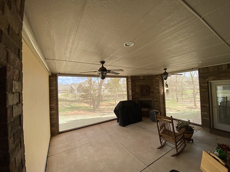 Covered patio with ceiling fans, brick fireplace, rocking chair, potted plant, and large screened windows showing a backyard with trees and houses.