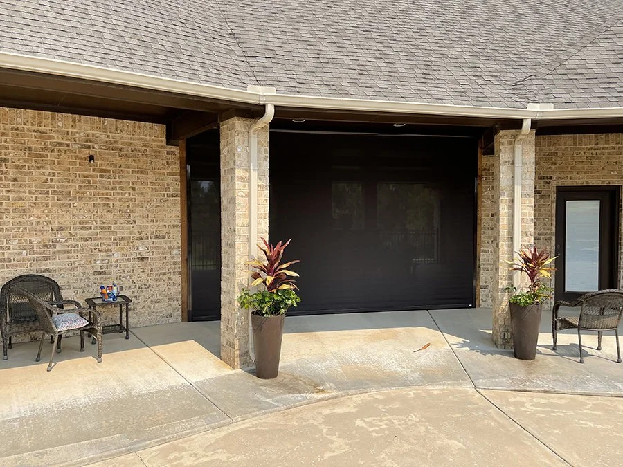 Patio area with two wicker chairs and a small table with toys on it, flanked by large potted plants with red and green leaves, on a concrete surface in front of a brick building with a black screen and a black door.