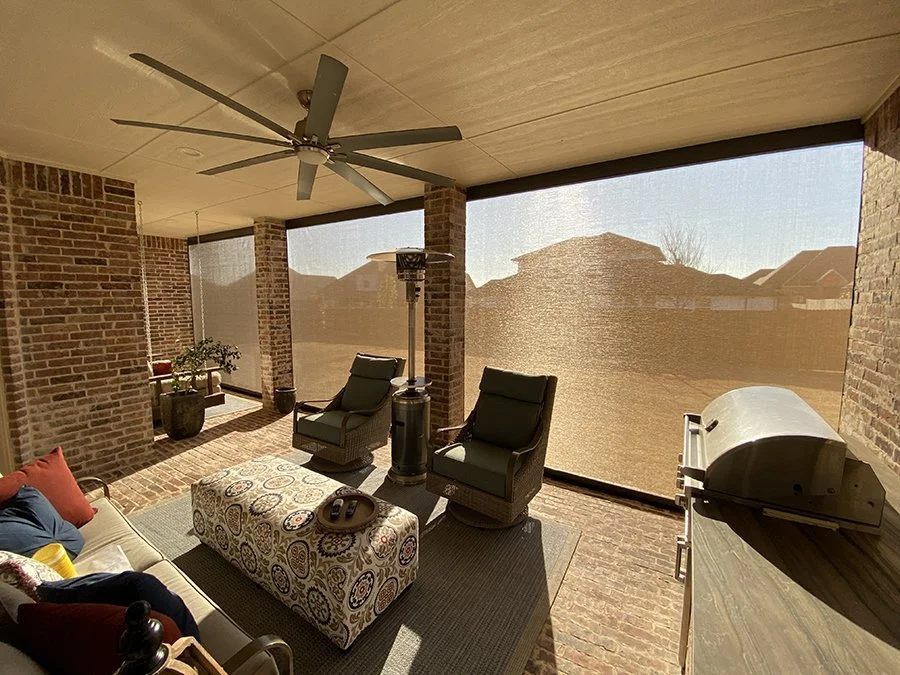 Sunlit screened porch with brick walls, ceiling fan, outdoor furniture, a patterned ottoman, a table, a grill, and a potted plant, with houses visible in the background.